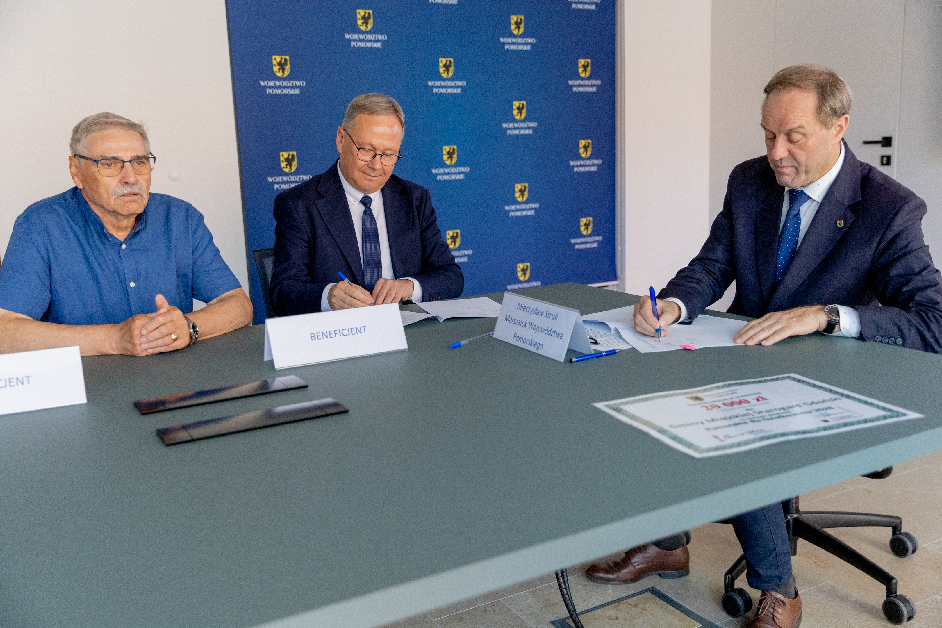 trzech mężczyzn przy stole dw&oacute;ch podpisuje dokumenty