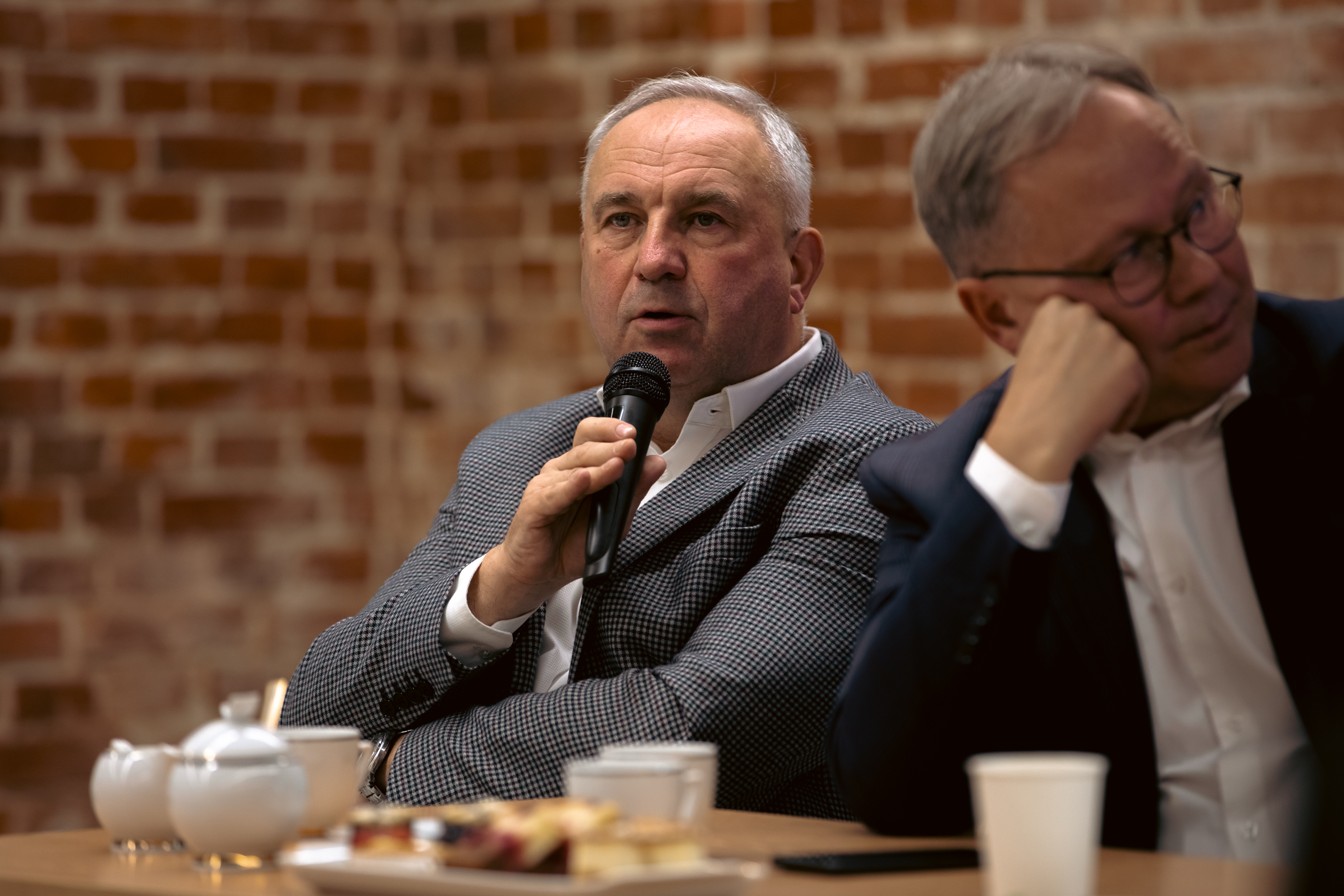 Dw&oacute;ch mężczyzn siedzi przy stole jeden z&nbsp;mikrofonem w&nbsp;dłoni przemawia