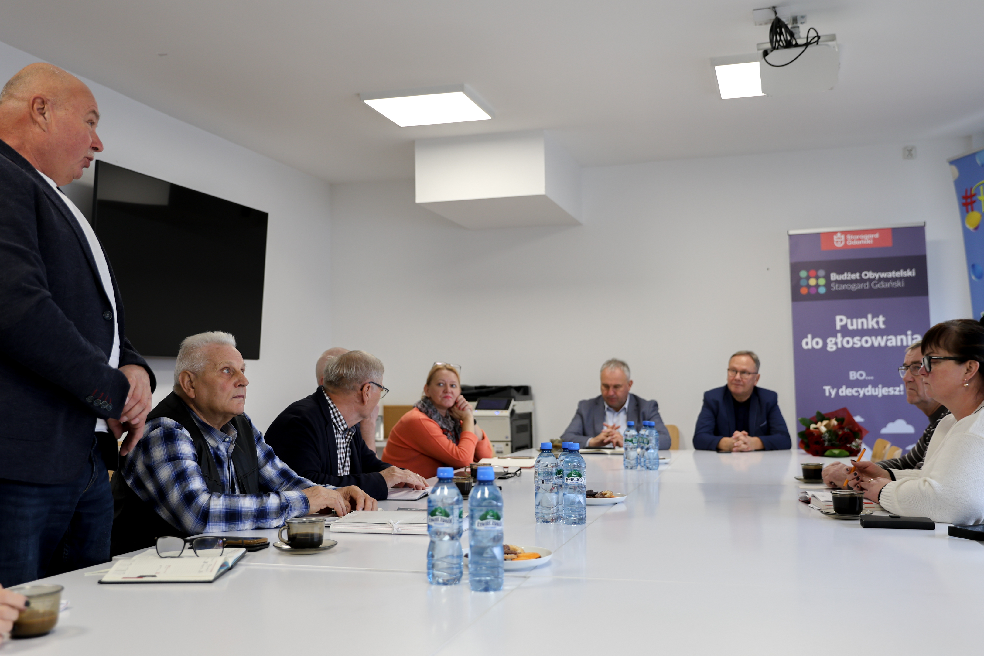 Ludzie siedzą wok&oacute;ł stołu jeden mężczyzna stoi