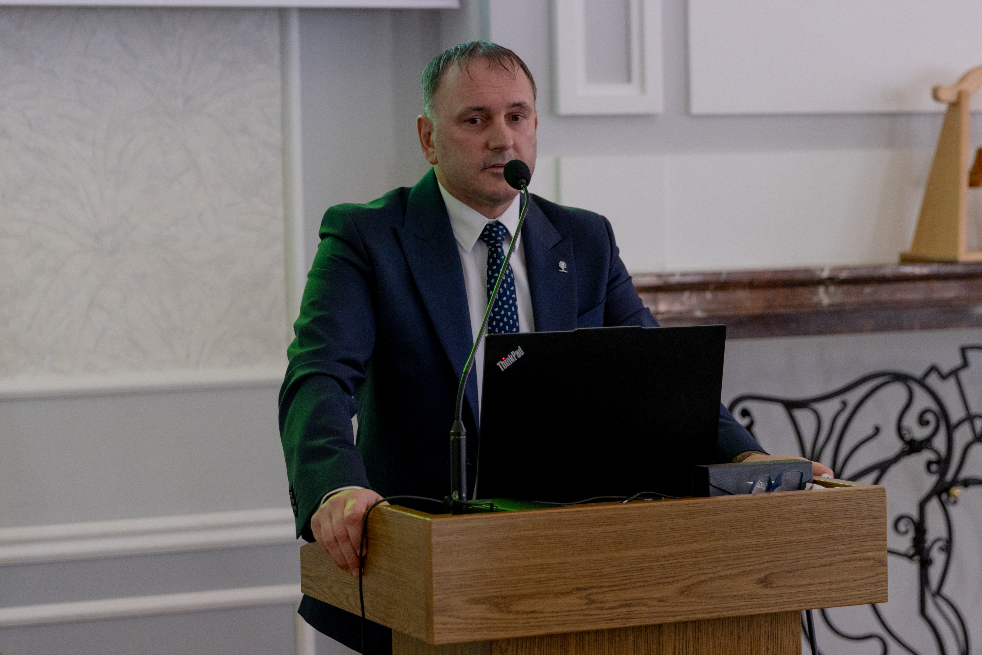 Mężczyzna w&nbsp;garniturze stoi przy m&oacute;wnicy i&nbsp;przemawia