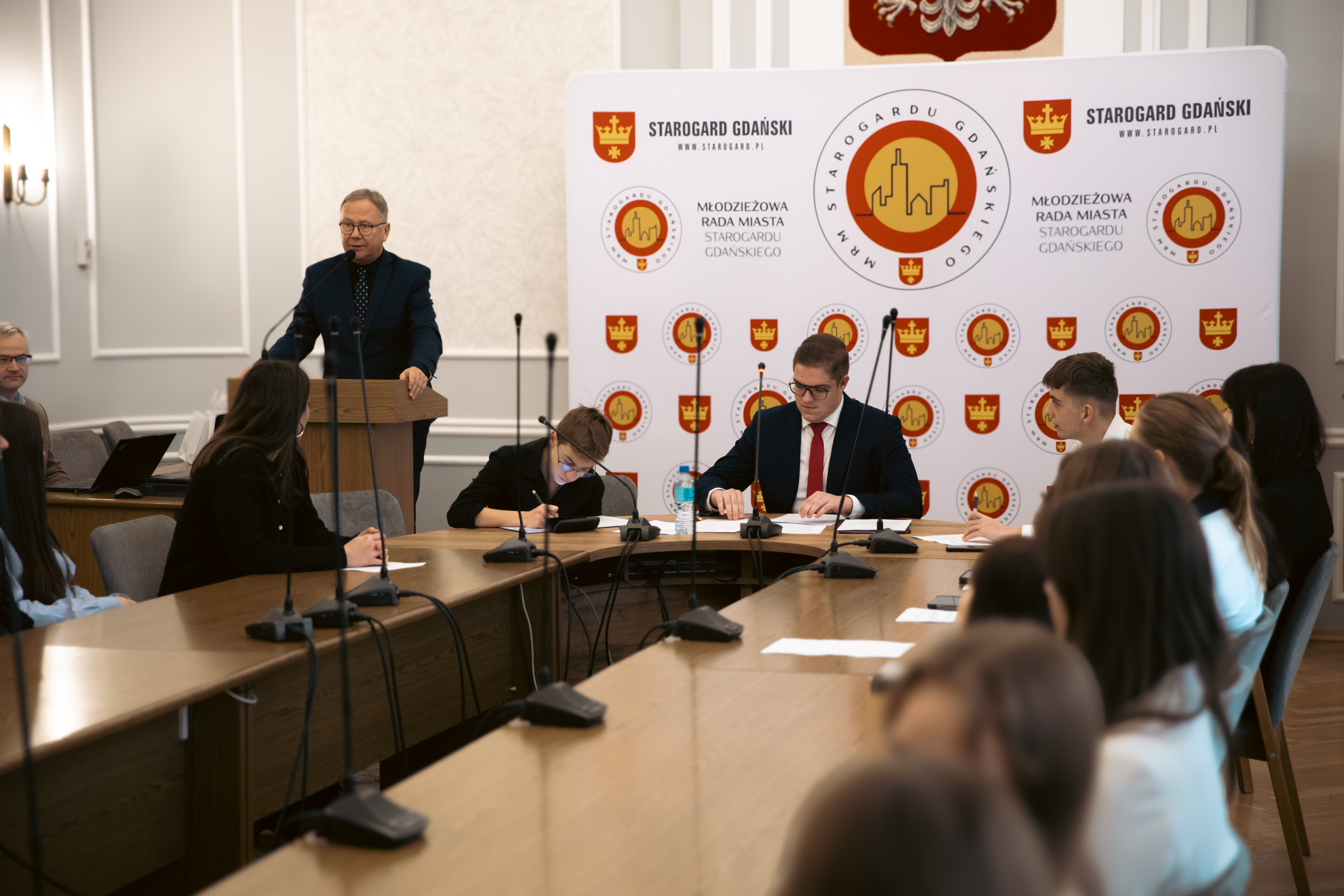 Młodziez siedzi przy owalnym stole i&nbsp;słucha przemawiającego na&nbsp;m&oacute;wnicy mężczyzny