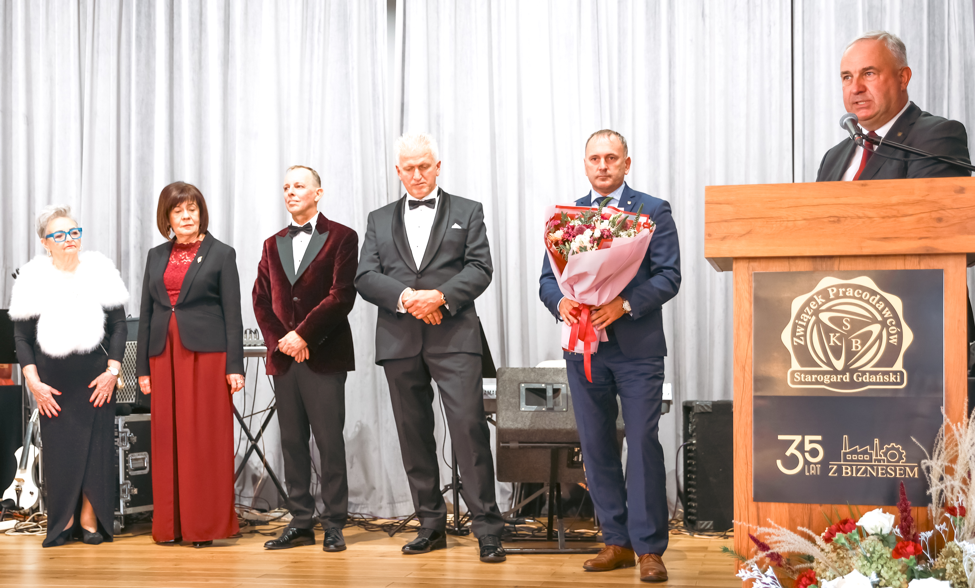 trzej mężczyźni i&nbsp;dwie kobiety elgancko ubrani stoją na&nbsp;scenie, czwarty mężczyzna na&nbsp;m&oacute;wnicy przemawia