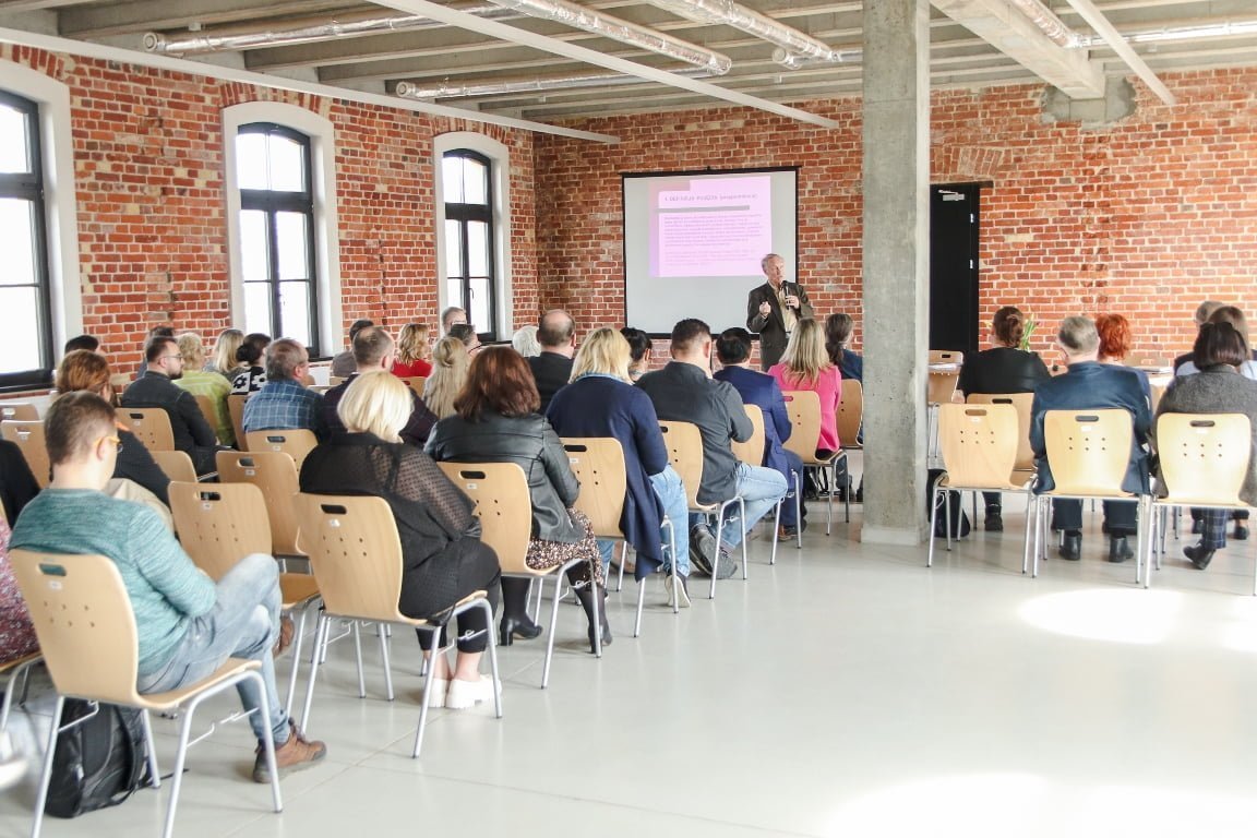 Na zdjeciu widać od tyłu uczetsników seminarium siedzących na krzesław, w tle widac ekran na którym prezentowane sa slajdy i wykładowcę z mikrofonem w reku.
