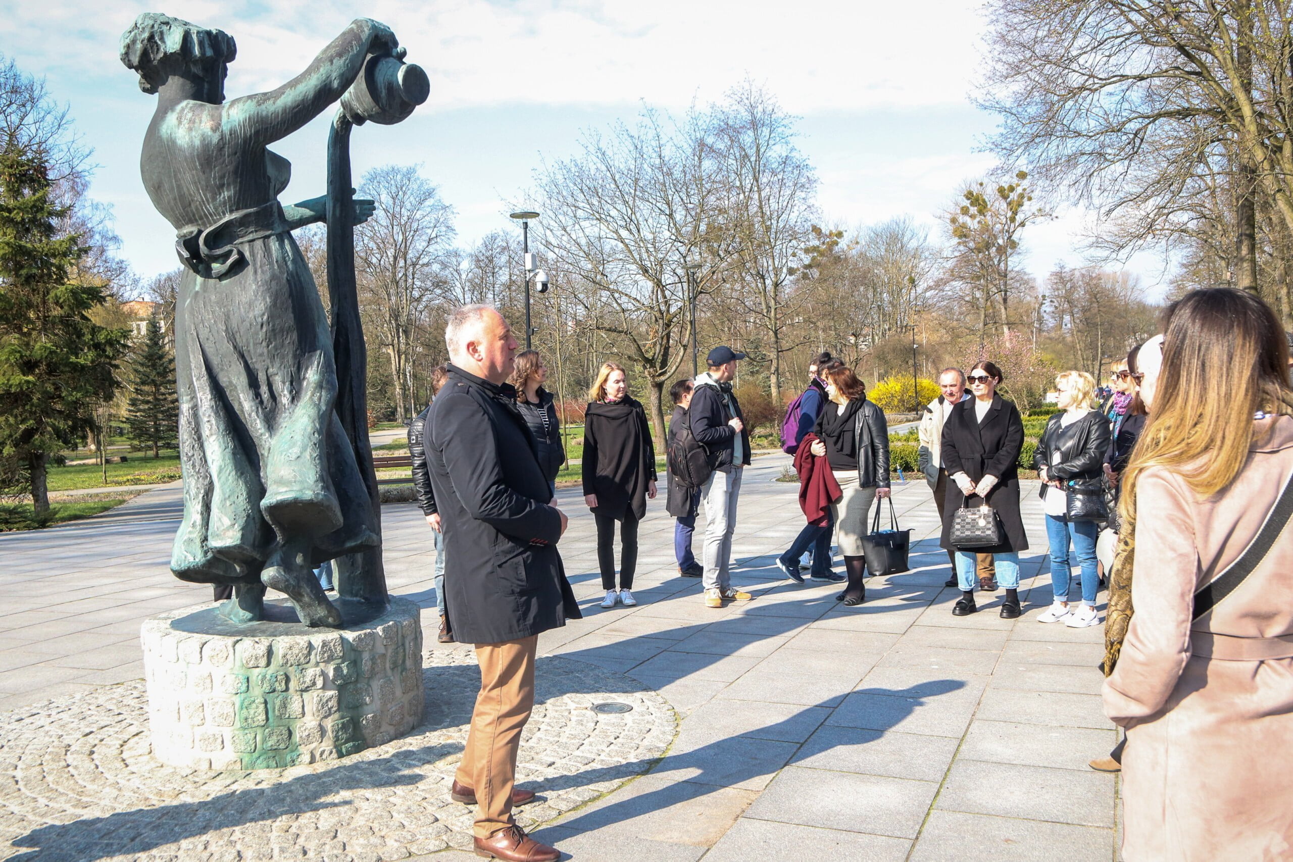 Park Miejski. Na pierwszym planie Wierzyczanka przy której stoi mezczyzna. Otaczają go ludzie , którzy słuchają tego co mówi. 