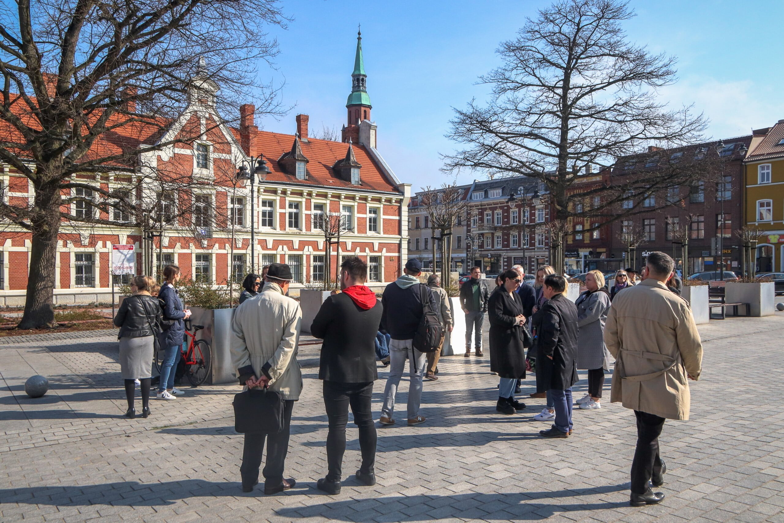 Rynek Miasta . Na placu przed budynkiem Ratusza stoi grupa ludzi , Rozmawiają.