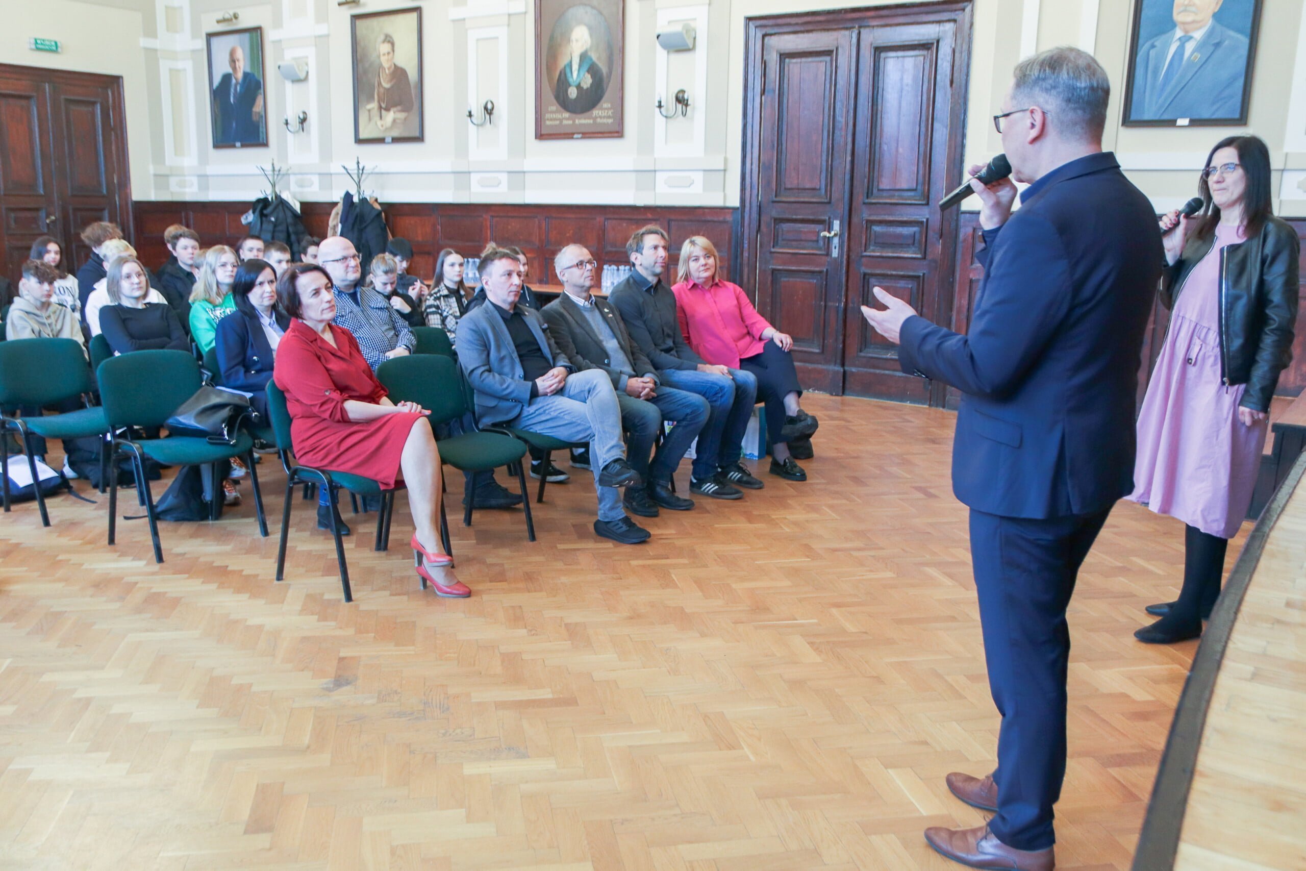 Na pierwszym froncie stoi mężczyzna z mikrofonem w reku i przemawia do siedzących na krzesłach osób. 
