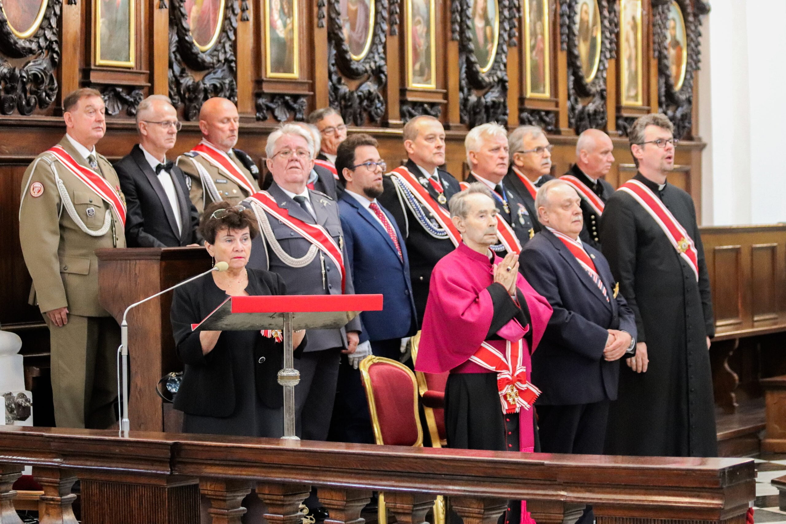 na zdjeciu wudać grupę starszych ludzi w elegancjich strojach z szarfami przewieszonymi przez bok. Przy mikrofonie stoi kobieta. Wszyscy znajdują się kościele