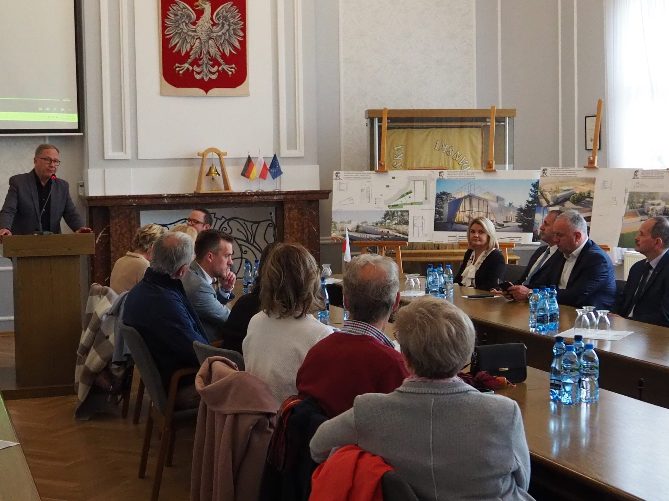 Ludzie siedza przy stole i słuchają. Mężczyzna przy mównicy stoi i przemawia. 
