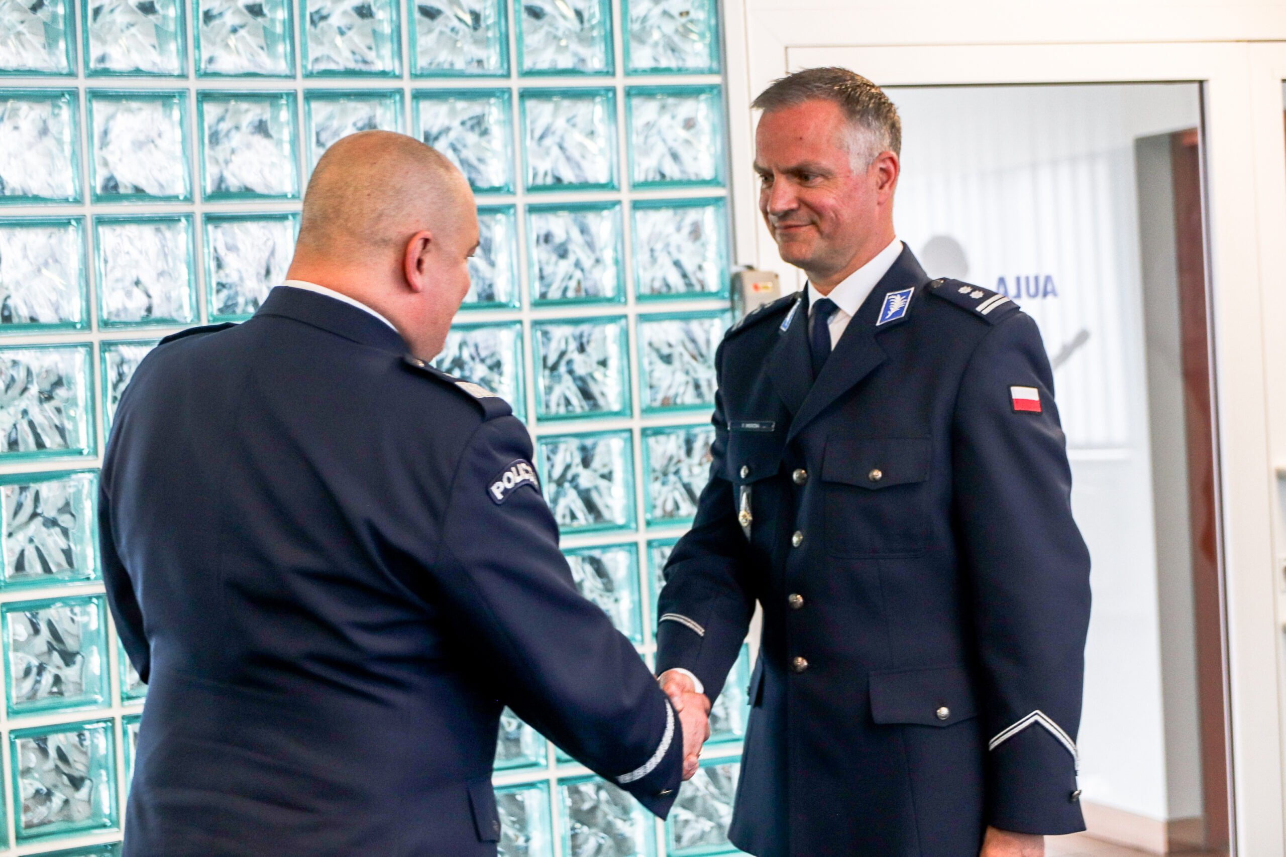 Dwóch mężczyzn w mundurach policyjnych stoi naprezciw siebie. Jeden przekazuje drugiemu akt powołania