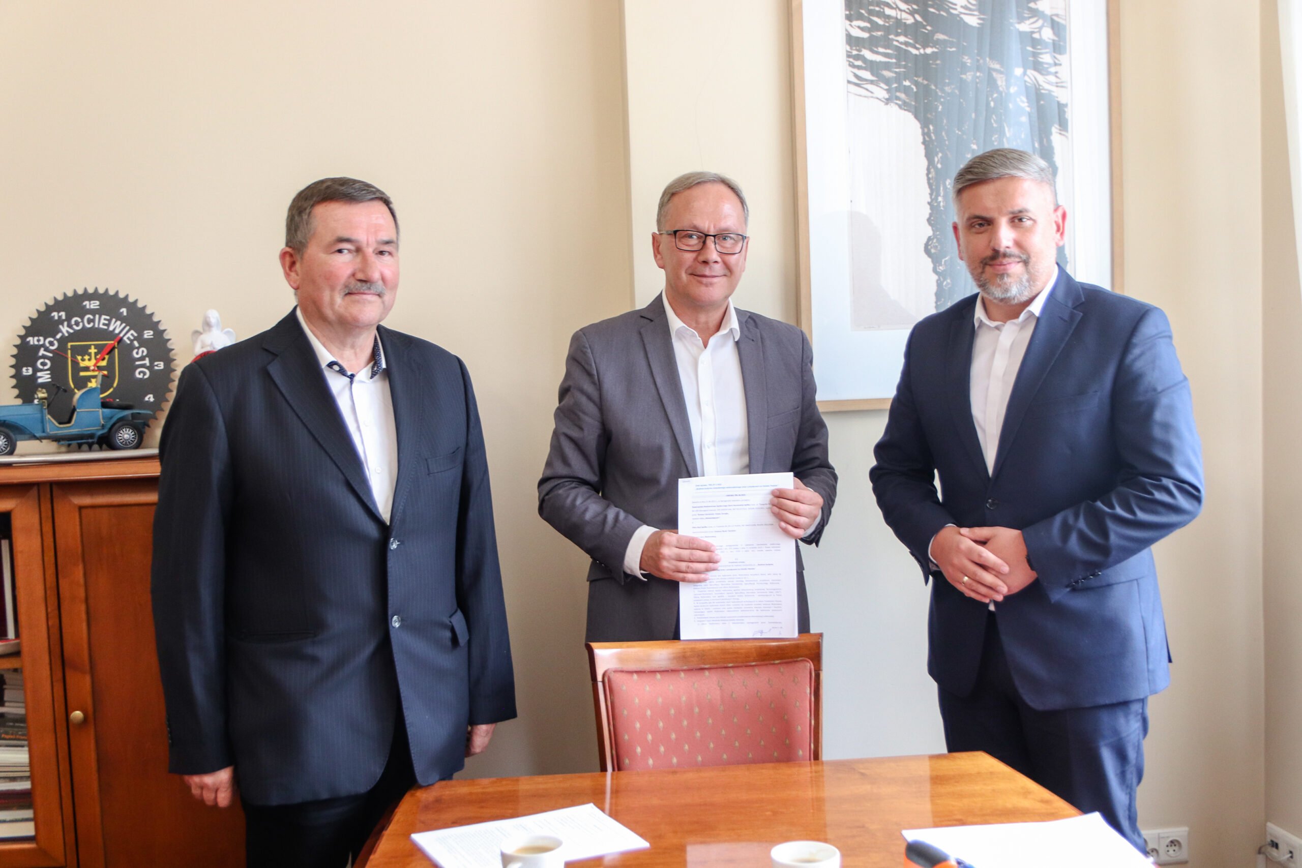Trzech mężczyzn. Srodkowy trzyma w dłoniach podpisaną umowę