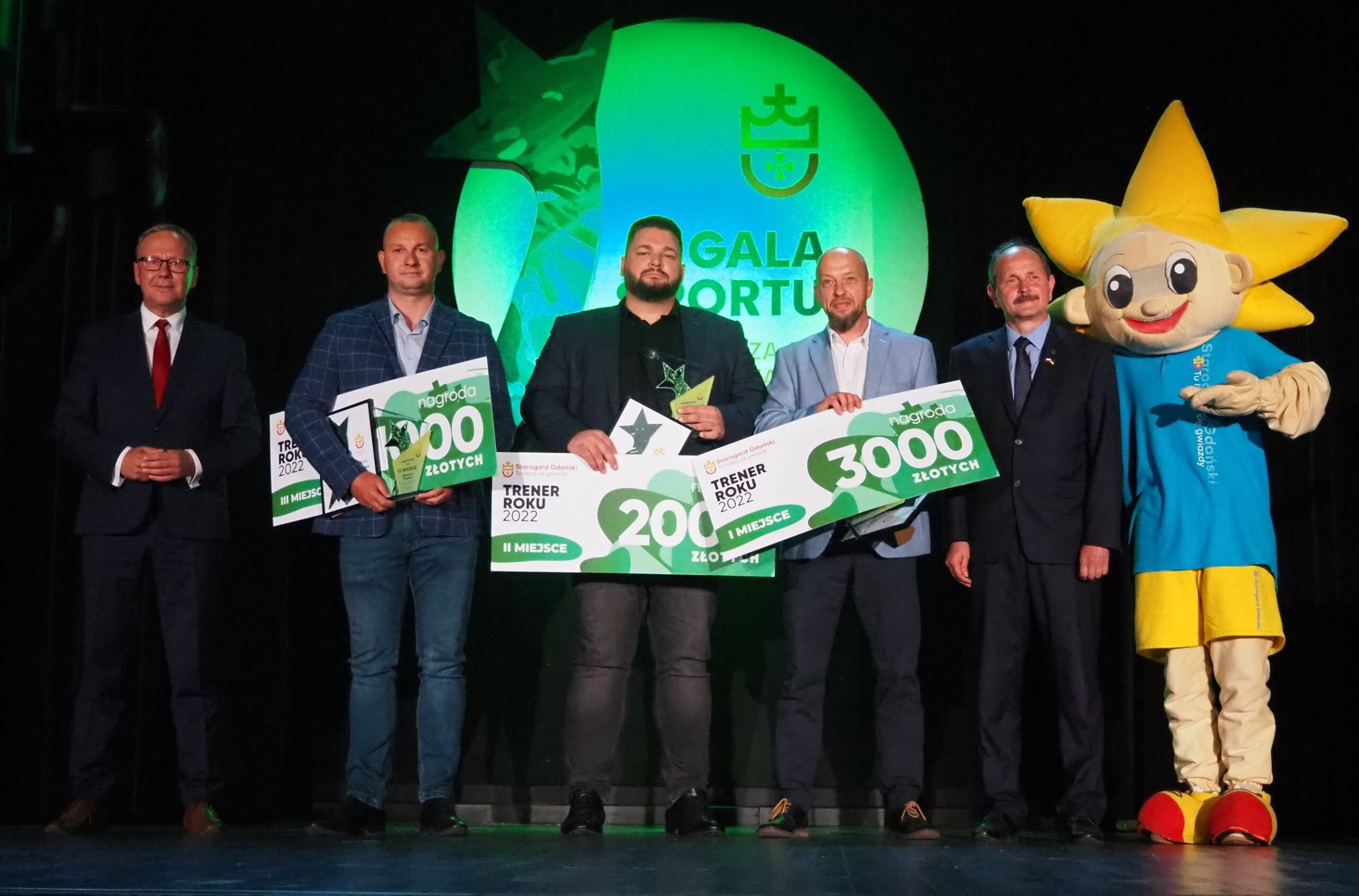Na scenie z Prezydentem Miasta Januszem Stankowiakiem, Przwodniczącą Rady Miasta Anną Benert i maskotką miejską Gwiazdkiem stoją Trenerzy Roku 2022: Marcin Drews (piłka nożna, KS Beniaminek 03 Starogard Gdański, III miejsce), Piotr Kamiński (piłka siatkowa, SKFiS KaEmKa Starogard Gdański, II miejsce) i Krzysztof Piątkowski (pika koszykowa, SKS Starogard Gdański, I miejsce).