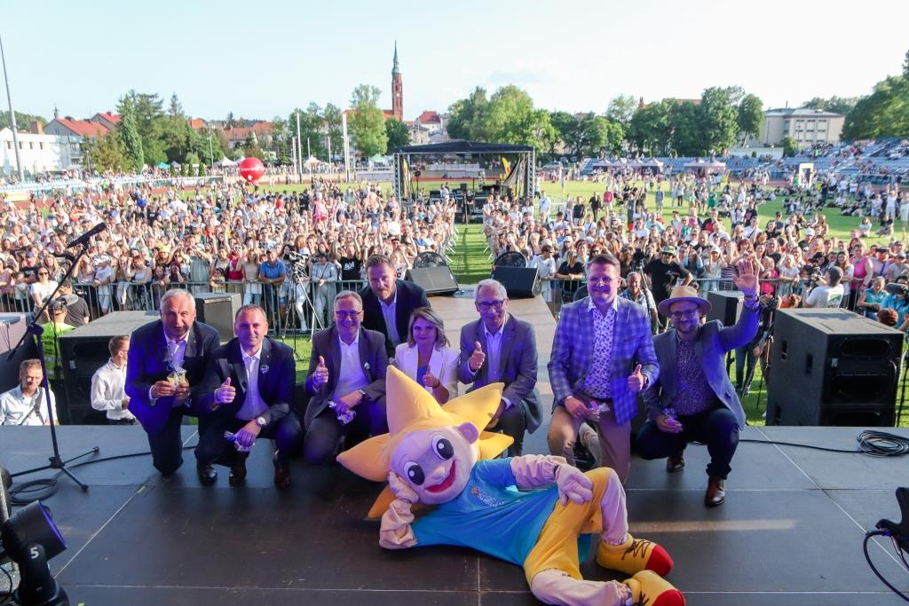 Władze miasta i zaproszeni goście pozują na scenie do pamiątkowego zdjęcia. Towarzyszy im maskotka miejska Gwiazdek. 