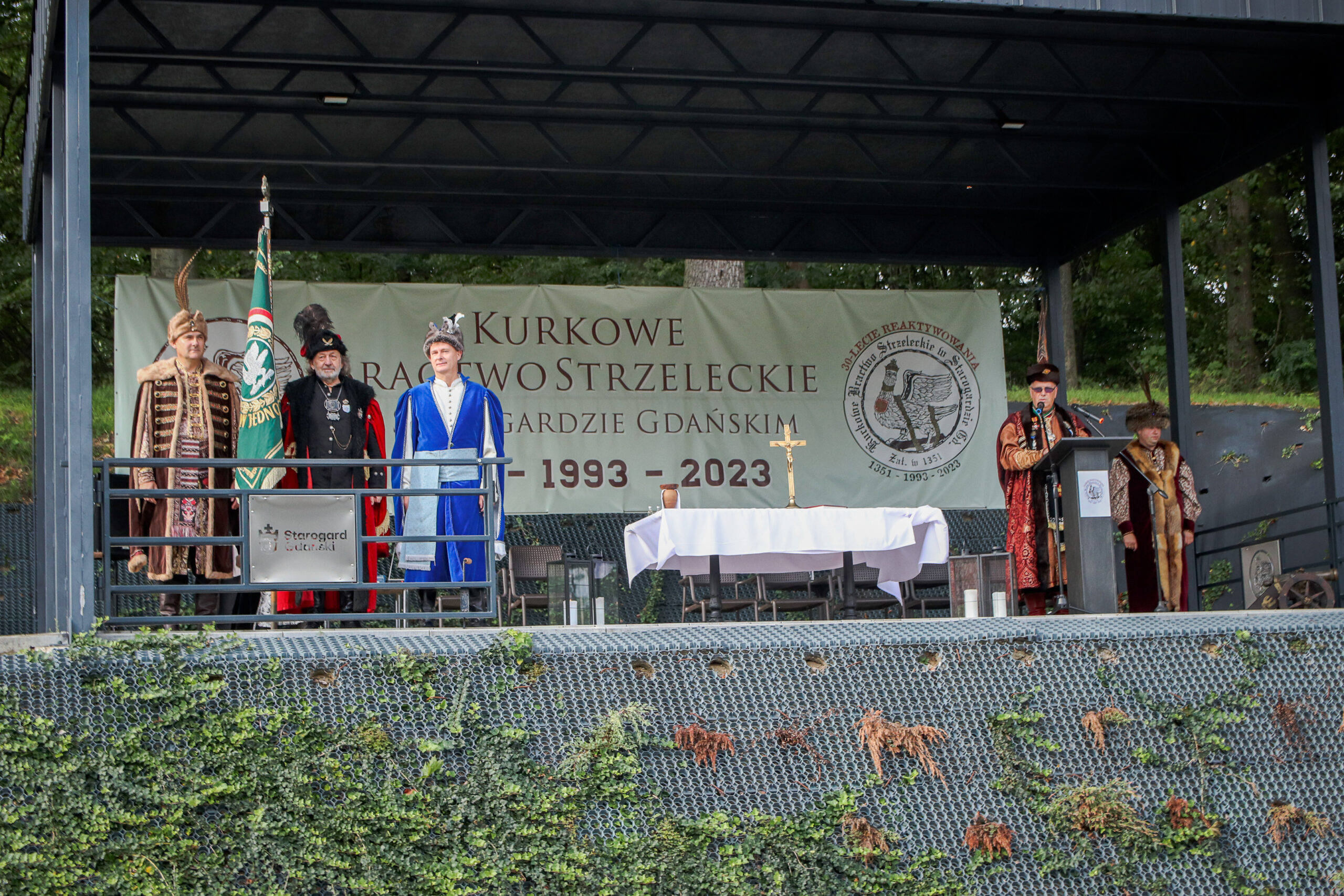 Na scenie stoi poczet sztandarowy. przy mikrowfonie stoi męzczyzna w kontuszu z czapką z piórami