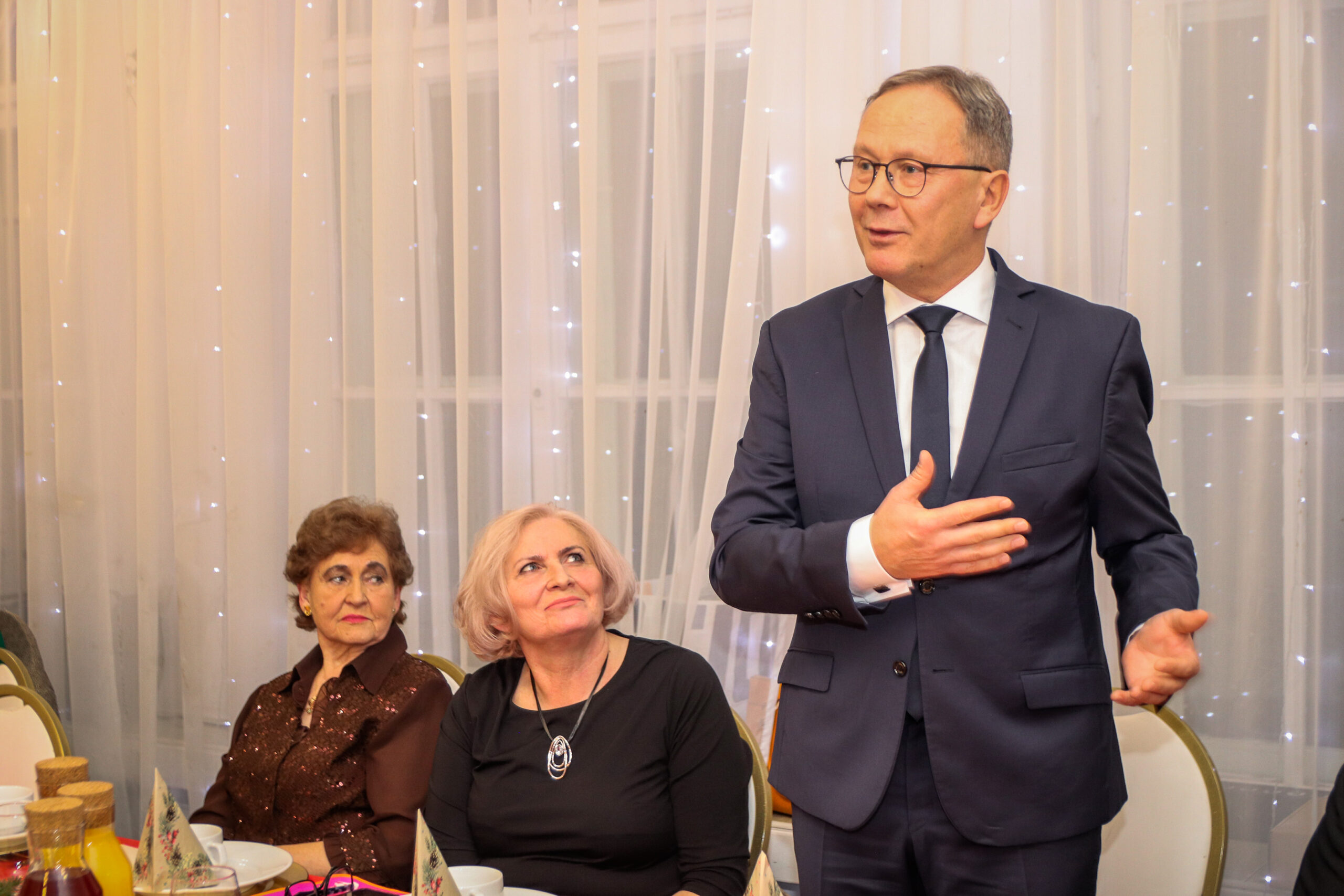 Mężczyzna stoi przy stole . Obok siedza dwie kobiety