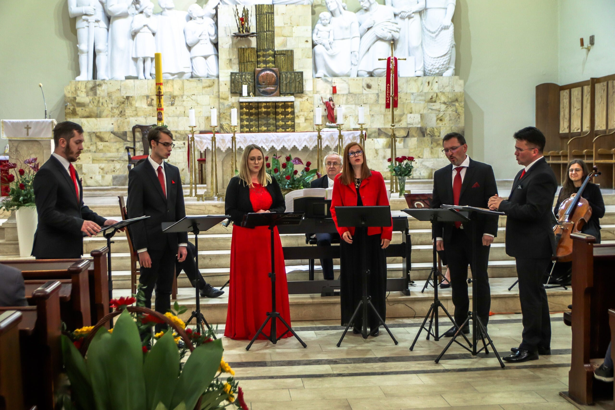 Czterech mężczyzn w garniiturach i dwie kobiety jedna w czerwonej druga w czranej sukience stoją na tle ołtarza śpiewają.